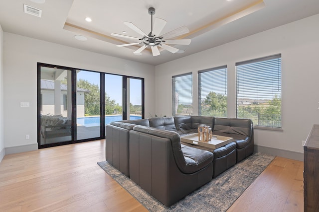 living room featuring ceiling fan, a raised ceiling, light hardwood / wood-style flooring, and a healthy amount of sunlight