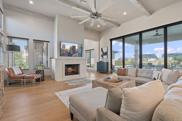 living room featuring beamed ceiling, a fireplace, light wood-type flooring, a high ceiling, and ceiling fan