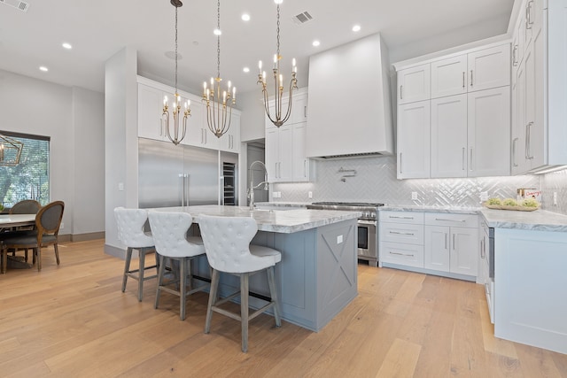 kitchen with custom exhaust hood, white cabinets, high end appliances, and an island with sink