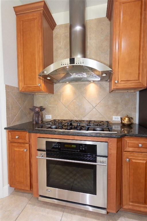 kitchen with wall chimney exhaust hood, dark stone countertops, light tile patterned floors, and appliances with stainless steel finishes