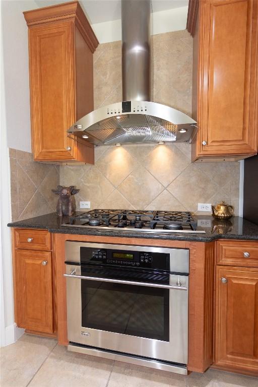 kitchen featuring appliances with stainless steel finishes, brown cabinetry, dark stone countertops, and wall chimney exhaust hood