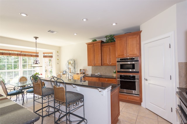 kitchen with stainless steel double oven, dark stone counters, decorative light fixtures, a kitchen island with sink, and light tile patterned floors