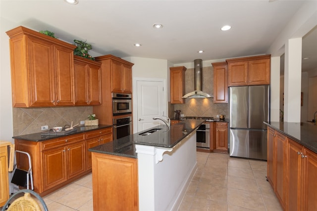 kitchen featuring appliances with stainless steel finishes, backsplash, wall chimney exhaust hood, sink, and a center island with sink