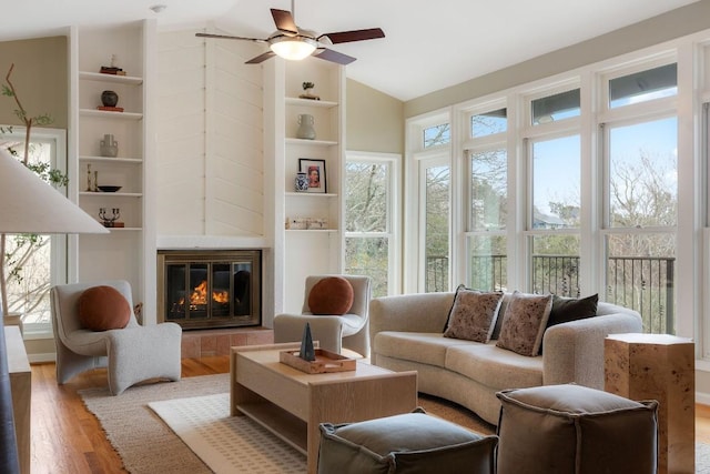 living room with built in features, a ceiling fan, a tiled fireplace, lofted ceiling, and wood finished floors