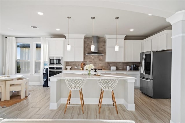 kitchen featuring pendant lighting, white cabinetry, stainless steel appliances, a center island with sink, and wall chimney range hood