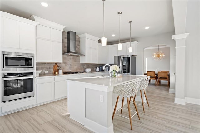 kitchen featuring tasteful backsplash, white cabinetry, a kitchen island with sink, stainless steel appliances, and wall chimney range hood