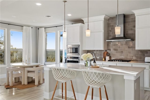 kitchen featuring wall chimney range hood, a kitchen island with sink, stainless steel appliances, a kitchen breakfast bar, and decorative light fixtures