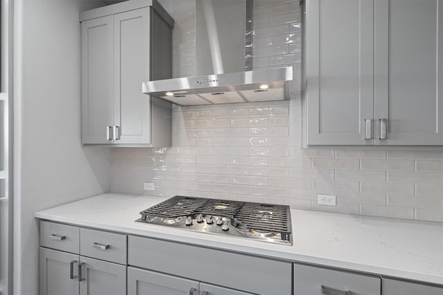 kitchen featuring stainless steel gas stovetop, backsplash, gray cabinets, light stone counters, and wall chimney range hood