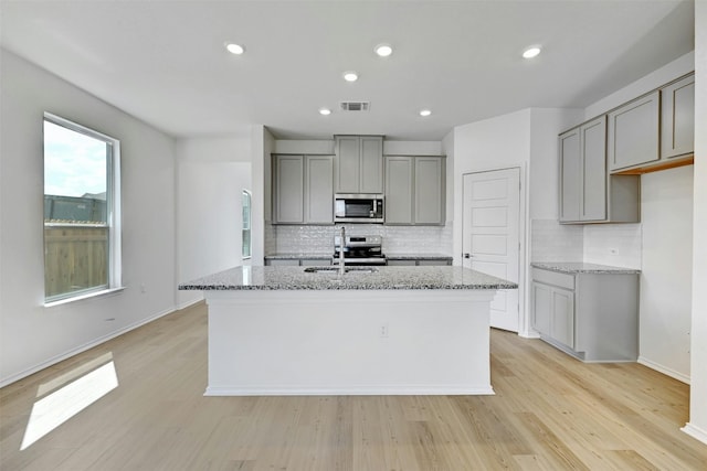 kitchen with sink, a center island with sink, light hardwood / wood-style flooring, stainless steel appliances, and light stone countertops