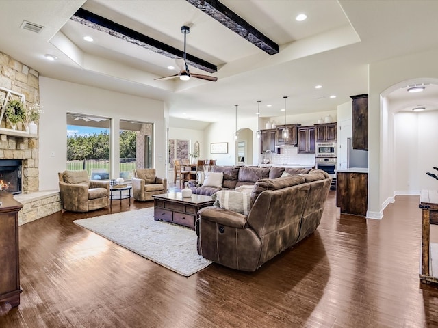 living room with dark wood-type flooring, beamed ceiling, ceiling fan, and a stone fireplace