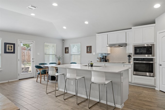 kitchen with built in microwave, white cabinetry, lofted ceiling, stainless steel oven, and a center island with sink