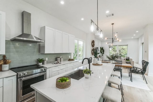 kitchen with sink, stainless steel range with electric cooktop, an island with sink, and wall chimney range hood