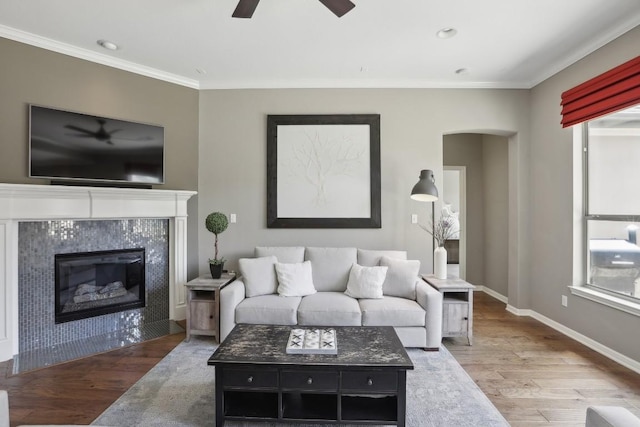 living room with wood finished floors, ceiling fan, ornamental molding, a tiled fireplace, and arched walkways