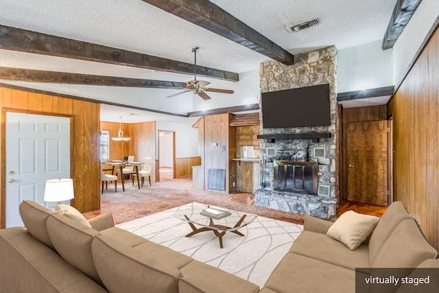 living room with ceiling fan, wood walls, beam ceiling, a stone fireplace, and light carpet