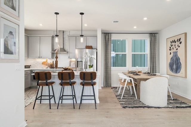 kitchen featuring stainless steel refrigerator with ice dispenser, pendant lighting, gray cabinets, wall chimney range hood, and light wood-type flooring