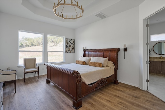 bedroom with a raised ceiling, ensuite bath, wood-type flooring, and a notable chandelier