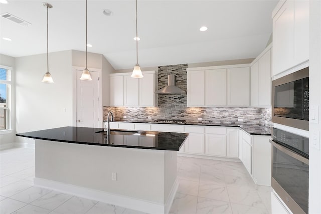 kitchen with white cabinets, hanging light fixtures, stainless steel appliances, an island with sink, and wall chimney exhaust hood