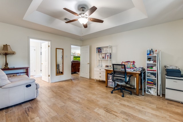 home office with light hardwood / wood-style flooring, ceiling fan, and a raised ceiling