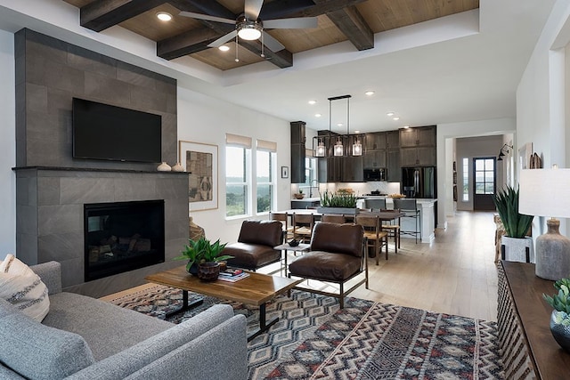 living room featuring a tiled fireplace, beam ceiling, wooden ceiling, light wood-type flooring, and ceiling fan