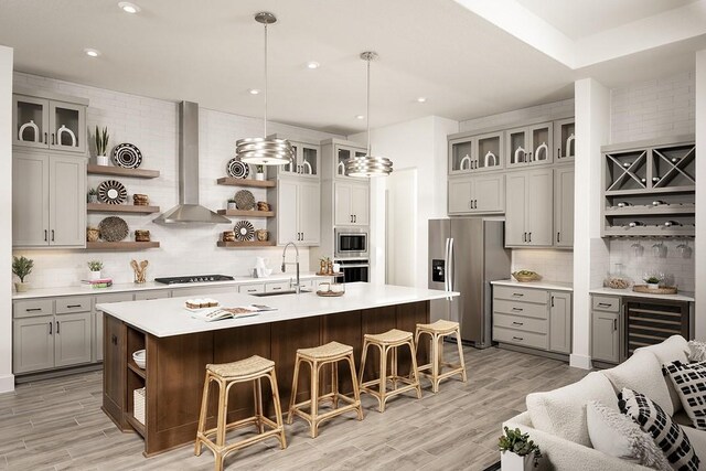 kitchen featuring wall chimney exhaust hood, stainless steel appliances, a center island with sink, pendant lighting, and light hardwood / wood-style floors