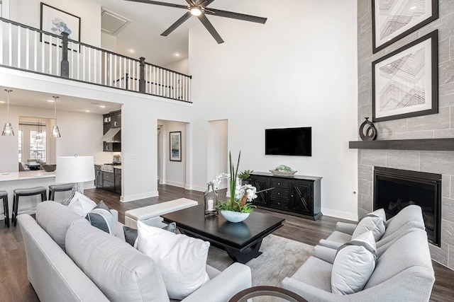 living room with ceiling fan, a fireplace, a towering ceiling, and dark wood-type flooring