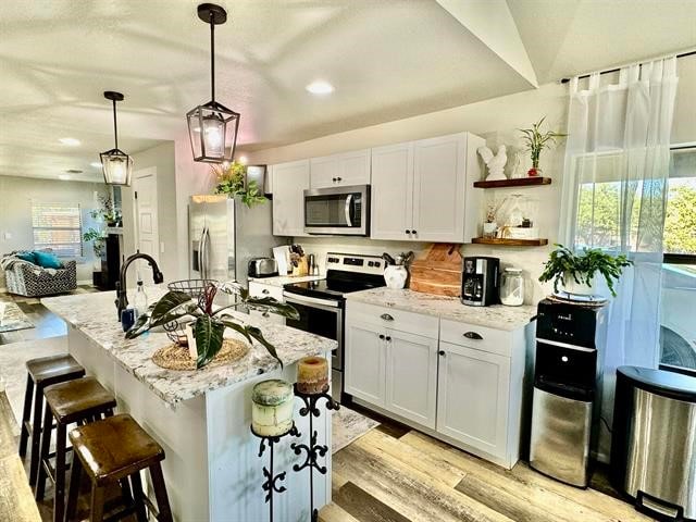 kitchen featuring pendant lighting, white cabinets, an island with sink, appliances with stainless steel finishes, and light stone counters