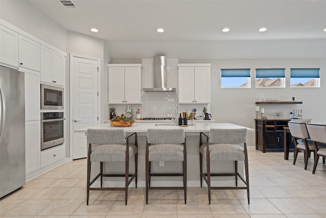 kitchen with a kitchen bar, an island with sink, wall chimney exhaust hood, appliances with stainless steel finishes, and white cabinets