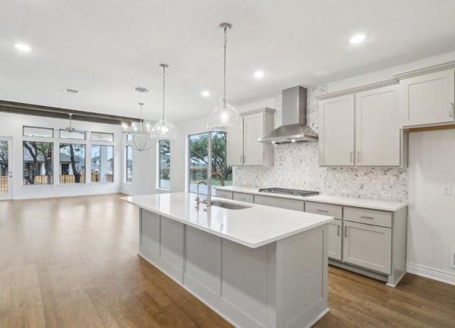 kitchen with wall chimney range hood, sink, a kitchen island with sink, backsplash, and decorative light fixtures