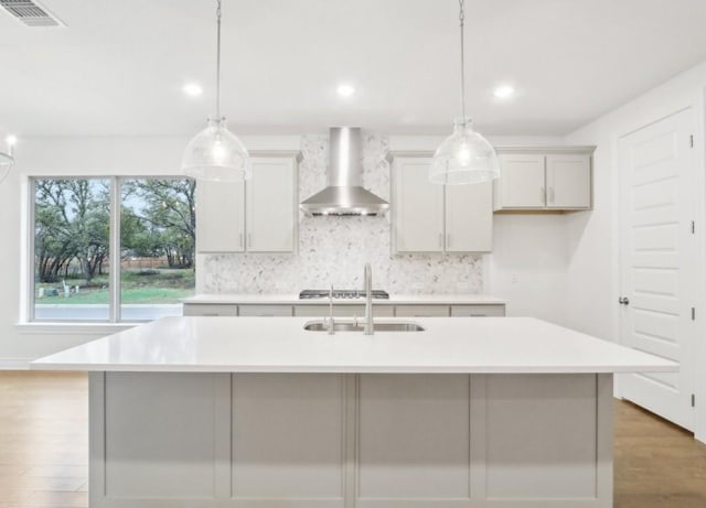 kitchen with hanging light fixtures, a kitchen island with sink, and wall chimney range hood