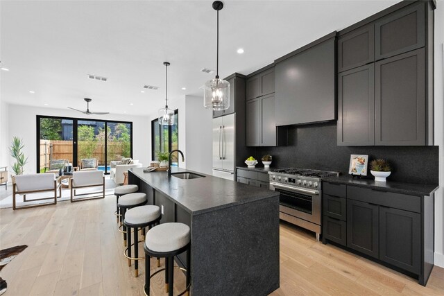 kitchen with a kitchen island with sink, sink, high end appliances, hanging light fixtures, and light hardwood / wood-style flooring