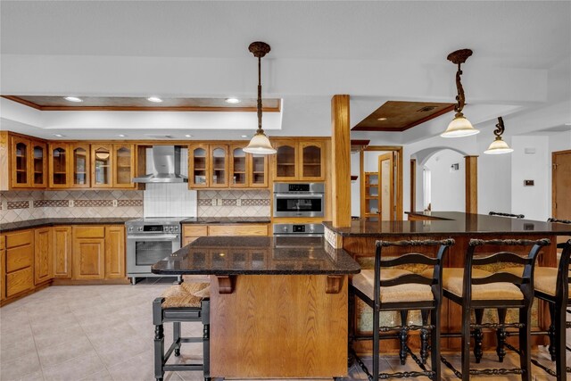 kitchen with appliances with stainless steel finishes, a kitchen breakfast bar, backsplash, decorative light fixtures, and wall chimney range hood