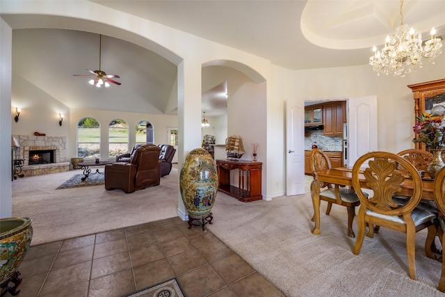 dining area with ceiling fan with notable chandelier, a stone fireplace, dark colored carpet, and high vaulted ceiling