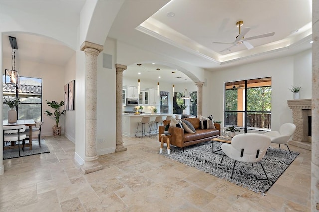 living area featuring ornate columns, ceiling fan, a raised ceiling, and a stone fireplace