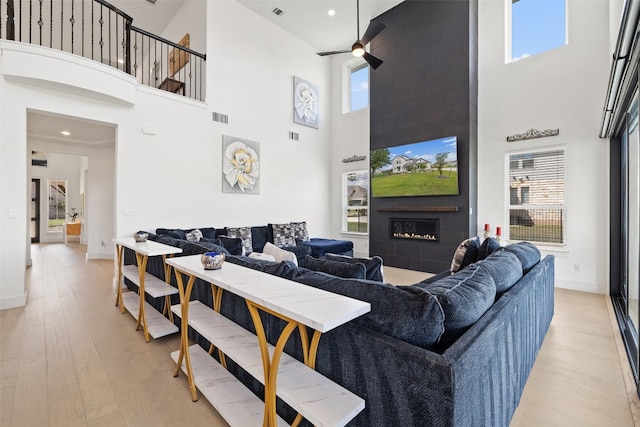 living room with ceiling fan, light wood-type flooring, a towering ceiling, a fireplace, and a healthy amount of sunlight