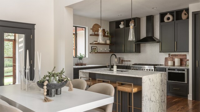 kitchen with wall chimney range hood, sink, appliances with stainless steel finishes, decorative light fixtures, and a center island with sink