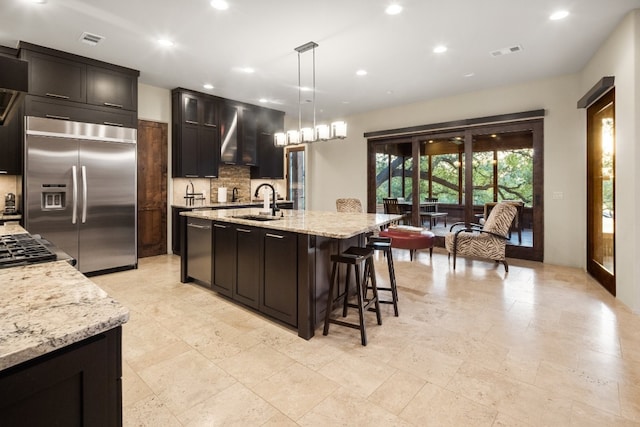 kitchen featuring sink, an island with sink, stainless steel built in fridge, decorative light fixtures, and light stone countertops