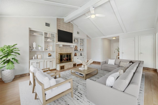 living room with light hardwood / wood-style floors, built in shelves, a brick fireplace, vaulted ceiling with beams, and ceiling fan