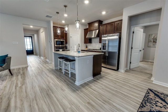 kitchen featuring light stone counters, a kitchen island with sink, ventilation hood, appliances with stainless steel finishes, and decorative light fixtures