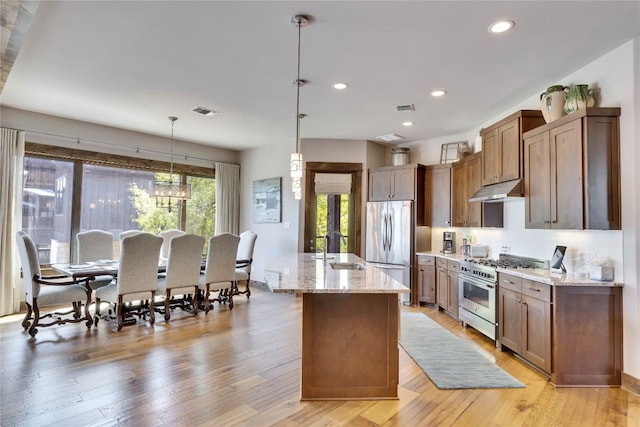 kitchen with appliances with stainless steel finishes, hanging light fixtures, light hardwood / wood-style floors, an island with sink, and light stone counters