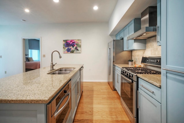 kitchen with light wood-type flooring, a kitchen island with sink, sink, stainless steel gas range oven, and wall chimney range hood