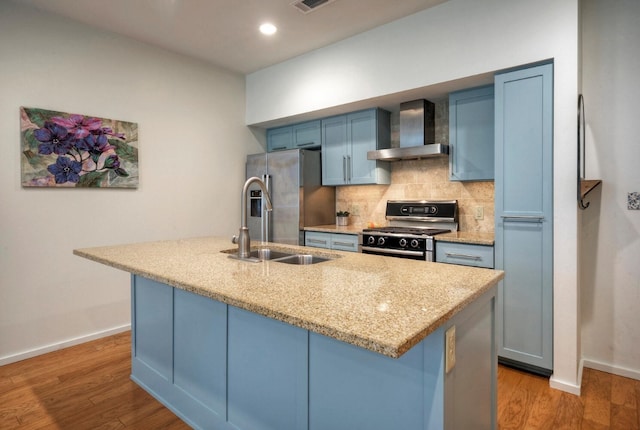 kitchen with light hardwood / wood-style floors, wall chimney exhaust hood, blue cabinetry, stainless steel appliances, and a kitchen island with sink