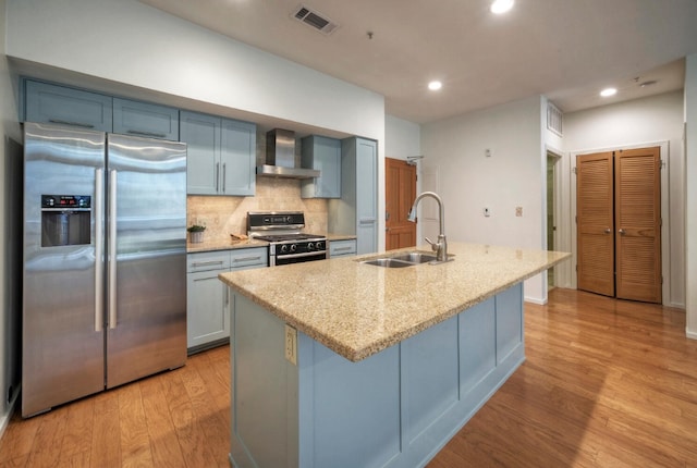 kitchen featuring sink, a center island with sink, wall chimney range hood, appliances with stainless steel finishes, and light wood-type flooring