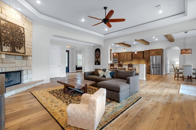 living room with ceiling fan, beam ceiling, ornamental molding, a stone fireplace, and light hardwood / wood-style flooring