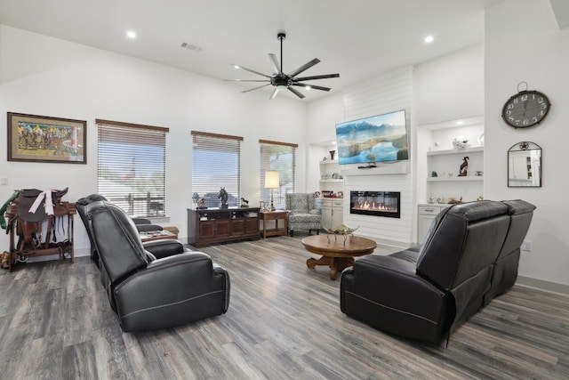 living room featuring a high ceiling, hardwood / wood-style floors, a large fireplace, ceiling fan, and built in features