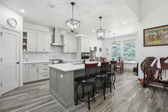 kitchen with dark wood-type flooring, a chandelier, pendant lighting, a kitchen island with sink, and wall chimney range hood