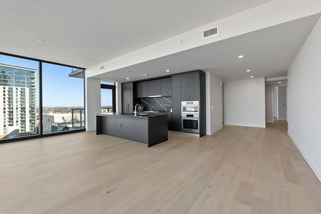 kitchen featuring backsplash, sink, light hardwood / wood-style flooring, an island with sink, and a wall of windows