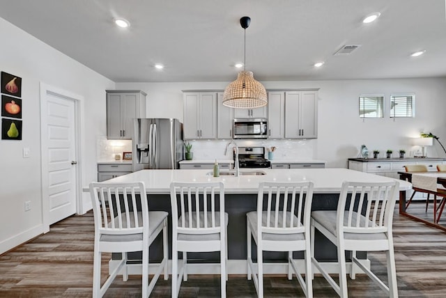 kitchen featuring dark wood-type flooring, stainless steel appliances, gray cabinets, a kitchen island with sink, and backsplash