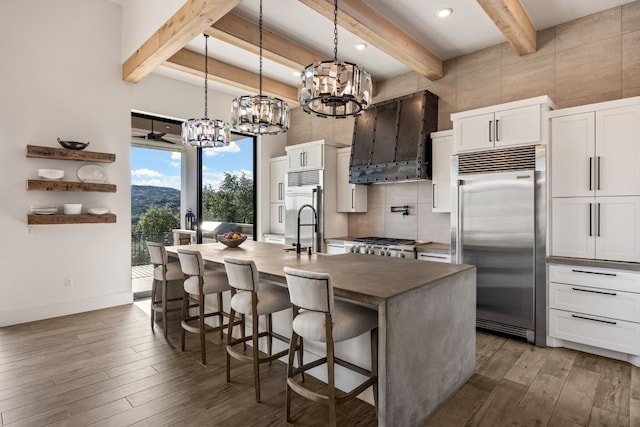 kitchen with premium range hood, a kitchen island with sink, built in refrigerator, and white cabinets