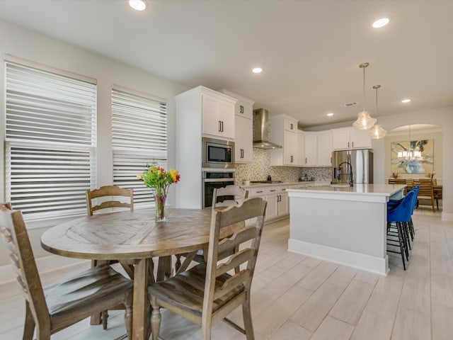 kitchen with a kitchen island with sink, white cabinets, wall chimney range hood, decorative light fixtures, and stainless steel appliances