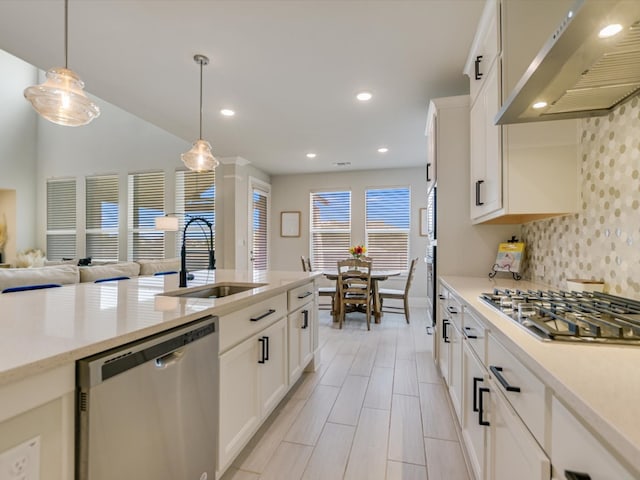 kitchen with wall chimney exhaust hood, stainless steel appliances, sink, white cabinets, and hanging light fixtures